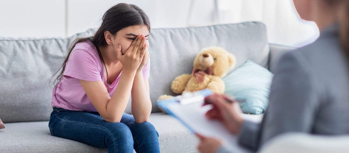 Depressed Indian teen girl having consultation with psychologist at office, covering face, talking about problems. Psychotherapist speaking with teenage patient at clinic, providing help and support